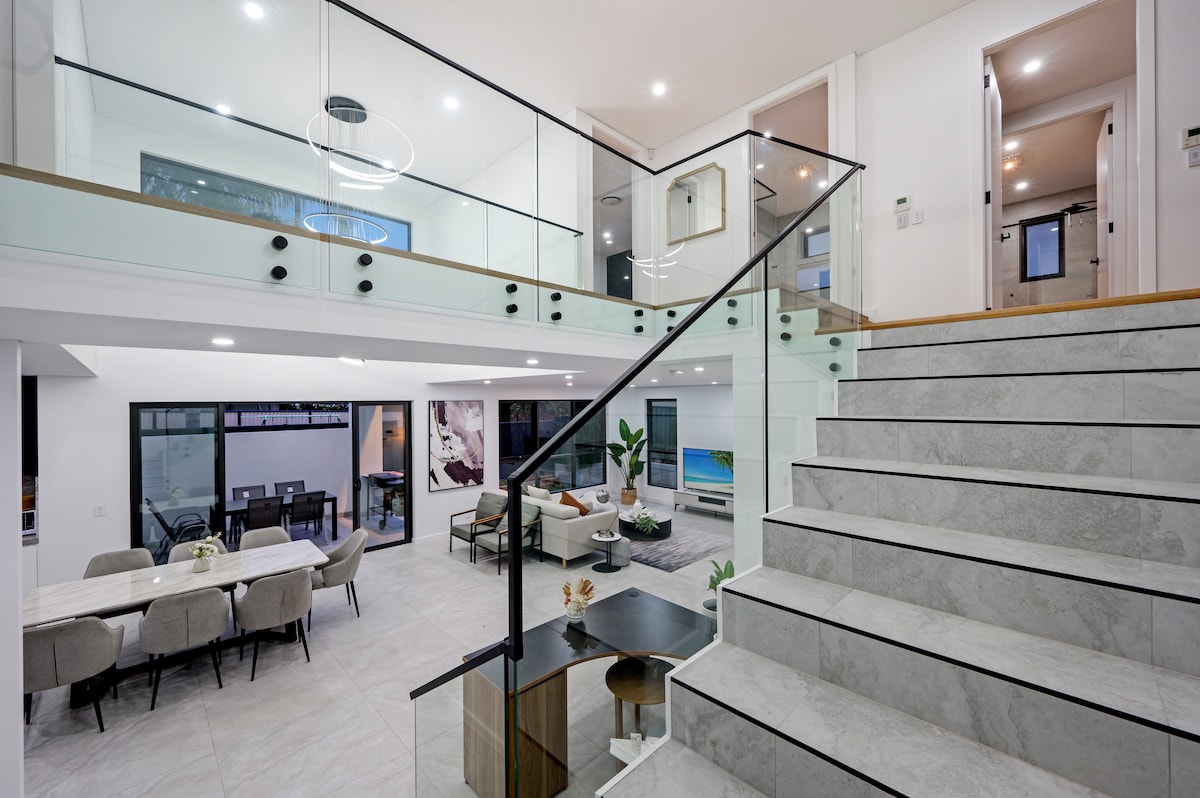 A modern staircase with a glass railing leads to an upper level. The open layout reveals a spacious living area with large windows and a dining table set for six. Soft lighting enhances the sleek furnishings and calming color palette throughout.