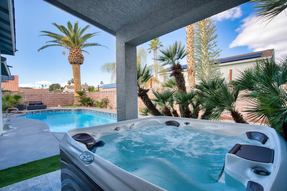 A view of a relaxing hot tub beside a shimmering pool is framed by palm trees and lush landscaping. The serene atmosphere is complemented by the bright blue sky and soft clouds, offering a tranquil outdoor setting.