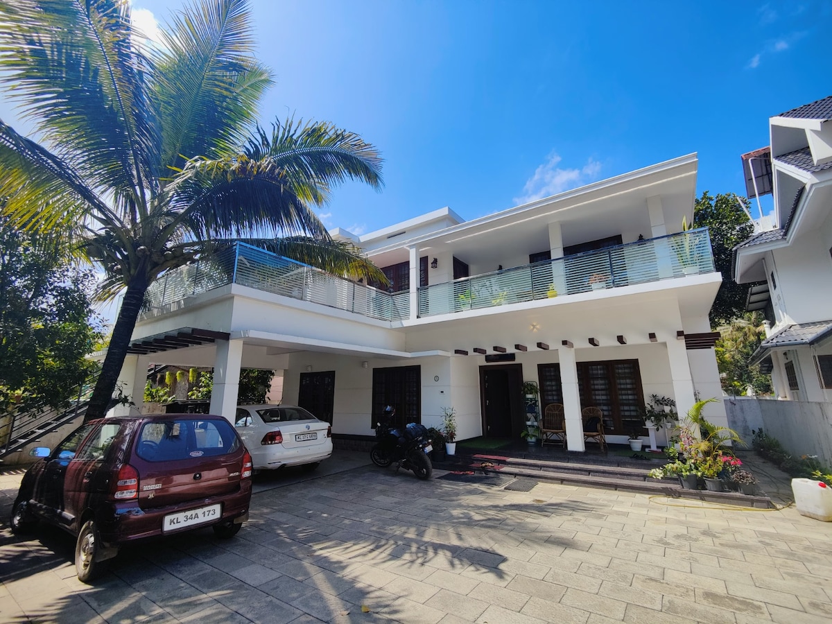 A modern two-story homestay is featured, surrounded by lush palm trees. A balcony extends from the upper floor, offering a view of the property. The driveway is paved and accommodates multiple vehicles, contributing to the overall welcoming appearance.