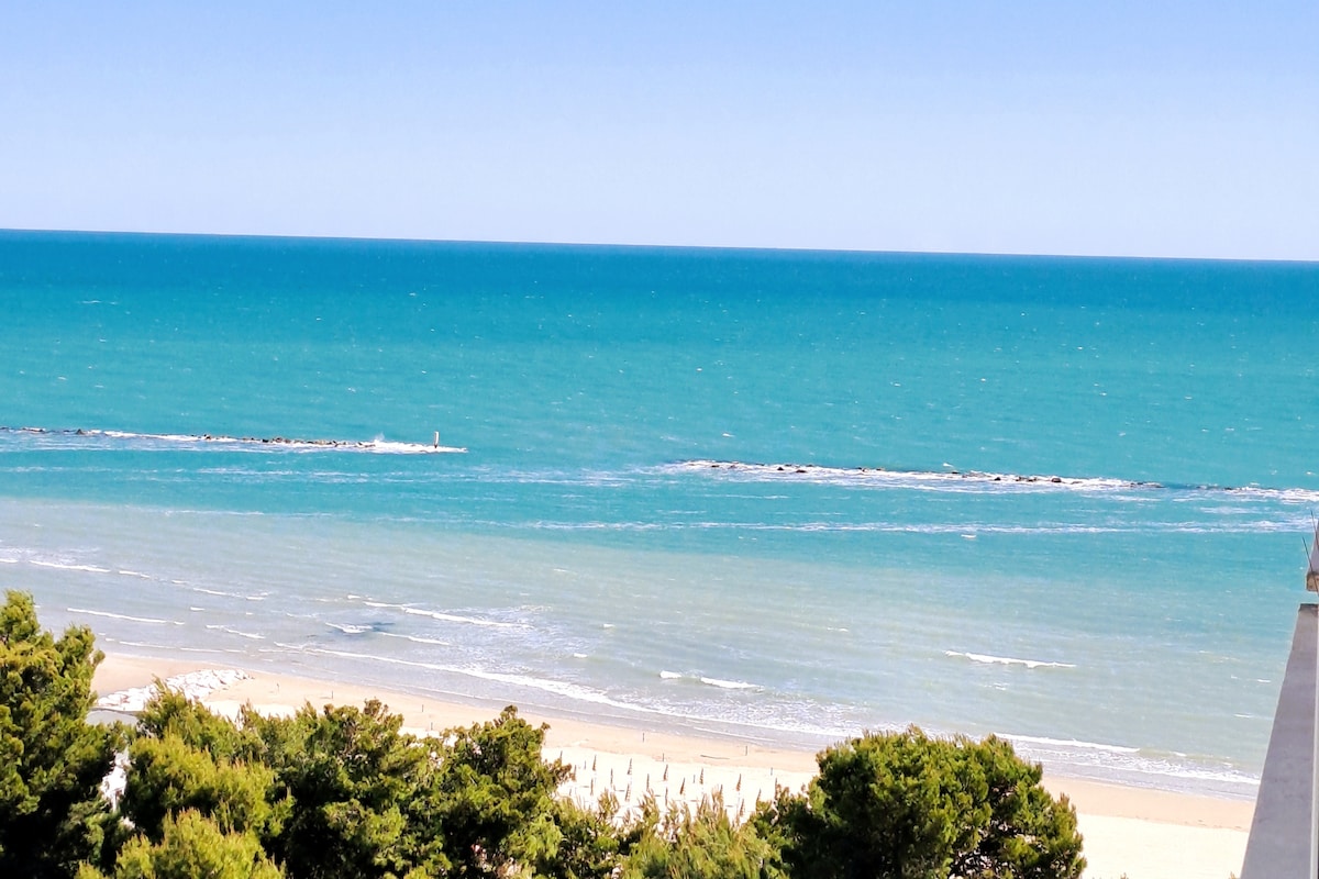 A panoramic view of the sea is presented, showcasing vibrant turquoise waters meeting the shore. Gentle waves can be seen lapping against a sandy beach adorned with umbrellas. The coastline is framed by lush greenery, enhancing the serene coastal atmosphere.