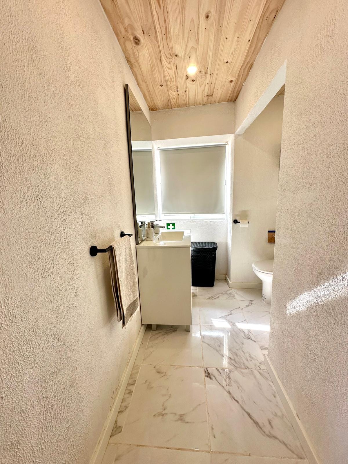 The bathroom features a modern design with a sleek white vanity and a large mirror mounted above. Natural light is provided by a window with shades. The floor is covered in elegant marble tiles, enhancing the space's clean and bright appearance.