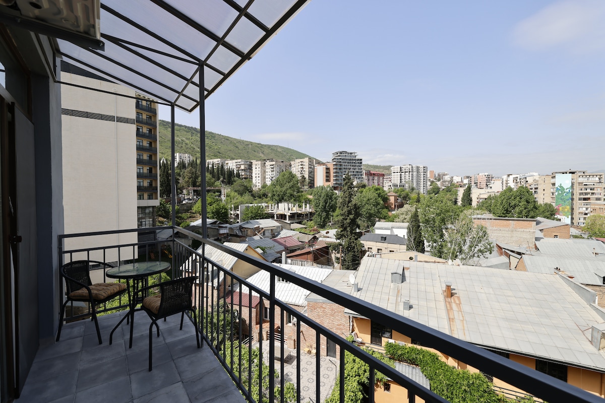 A balcony view overlooks a mix of urban and natural scenery, showcasing rooftops and greenery under a clear sky. A table with two chairs is positioned on the balcony, inviting relaxation while enjoying the surrounding vibrant neighborhood.