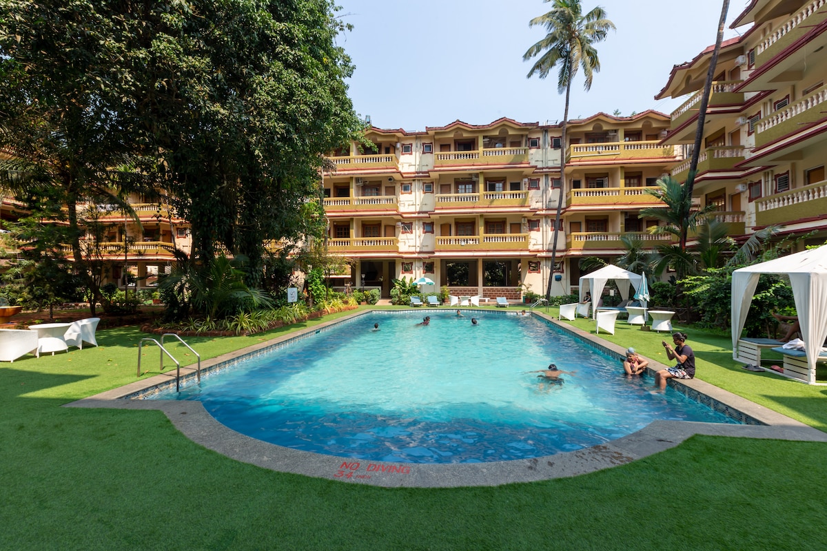 A refreshing swimming pool is set amidst lush greenery, surrounded by a well-maintained lawn. Several guests can be seen enjoying the water, while shaded cabanas provide areas for relaxation. The three-story building with balconies provides a backdrop to the tranquil scene.