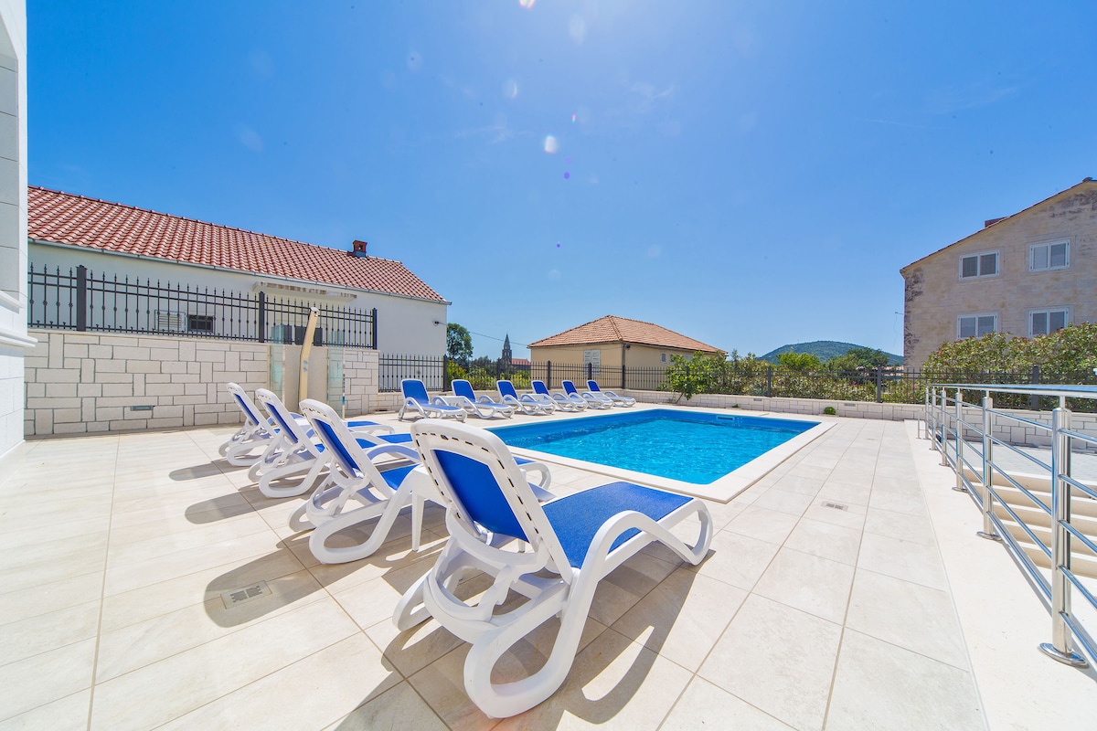 A well-maintained outdoor area features a shimmering pool surrounded by sun loungers with blue cushions. The patio is lined with neutral tiles, providing ample space for relaxation. Nearby, two residential buildings and greenery are visible under a clear blue sky.