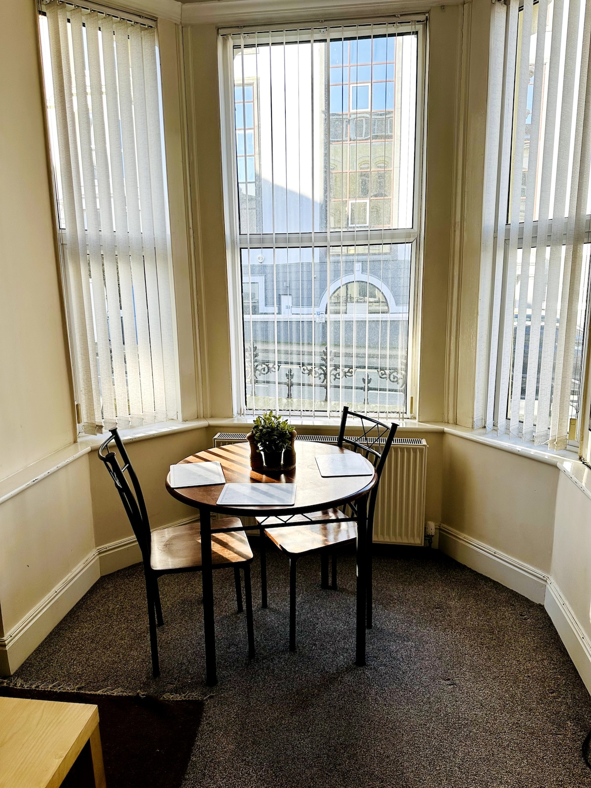 A small dining area includes a round wooden table surrounded by two chairs. Natural light floods in through large windows with vertical blinds, revealing views of the street outside. A small potted plant adds a touch of greenery to the table.