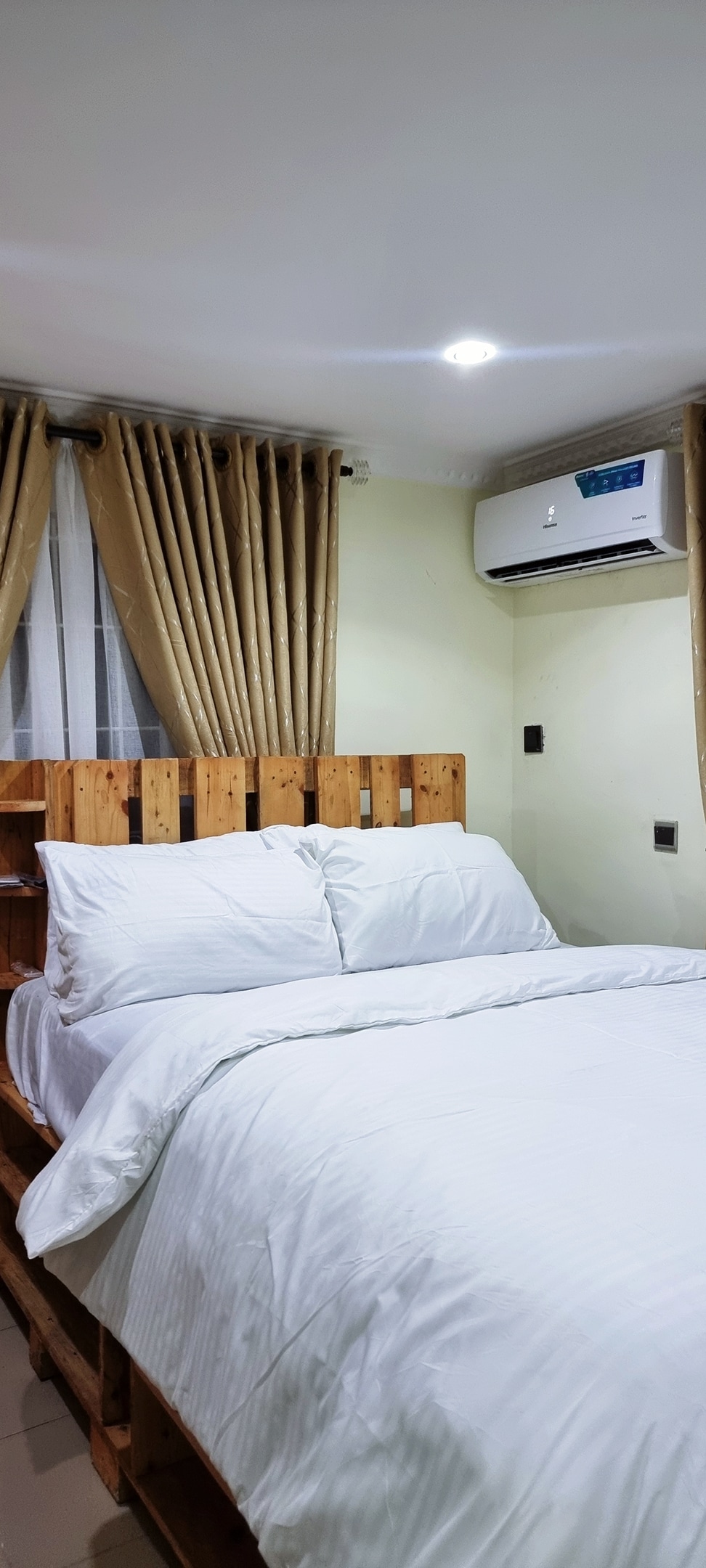A sleeping area features a neatly made bed with white linens and decorative pillows against a wooden headboard. Natural light enters through sheer curtains, contributing to the bright ambiance. An air conditioning unit is mounted on the wall for comfort.
