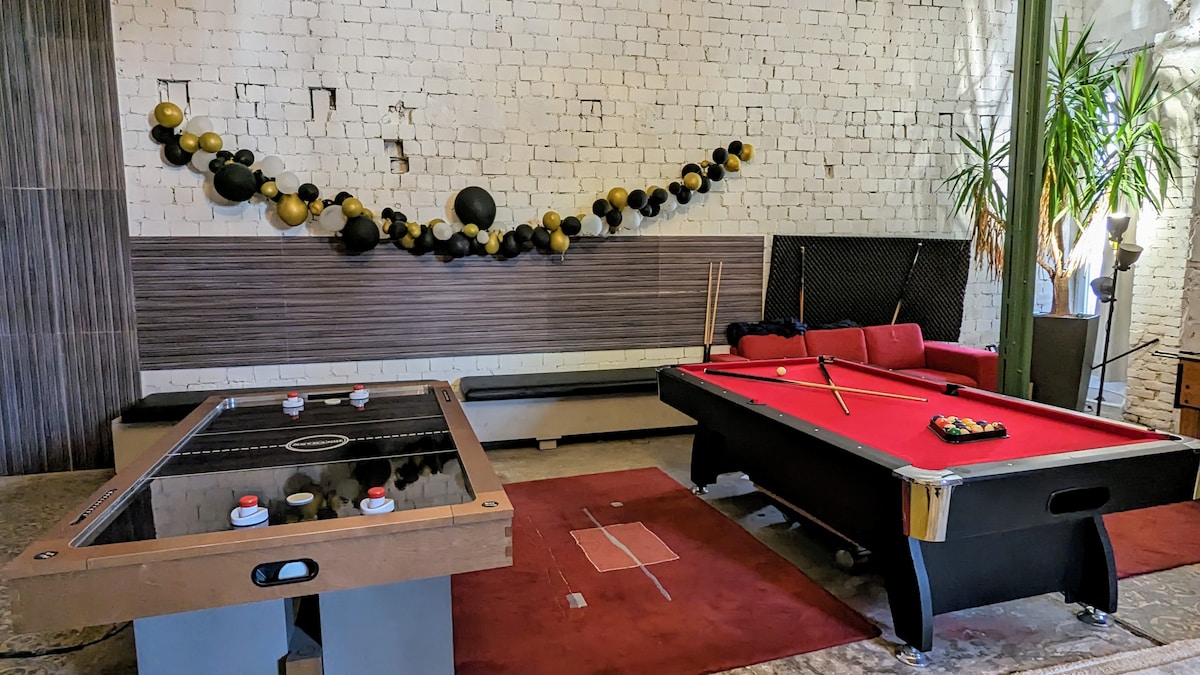 A recreational area is showcased, featuring a pool table and an air hockey table. A vibrant red seating area is positioned nearby, complemented by decorative elements on the wall. Natural light filters in, enhancing the spacious ambiance of the room.