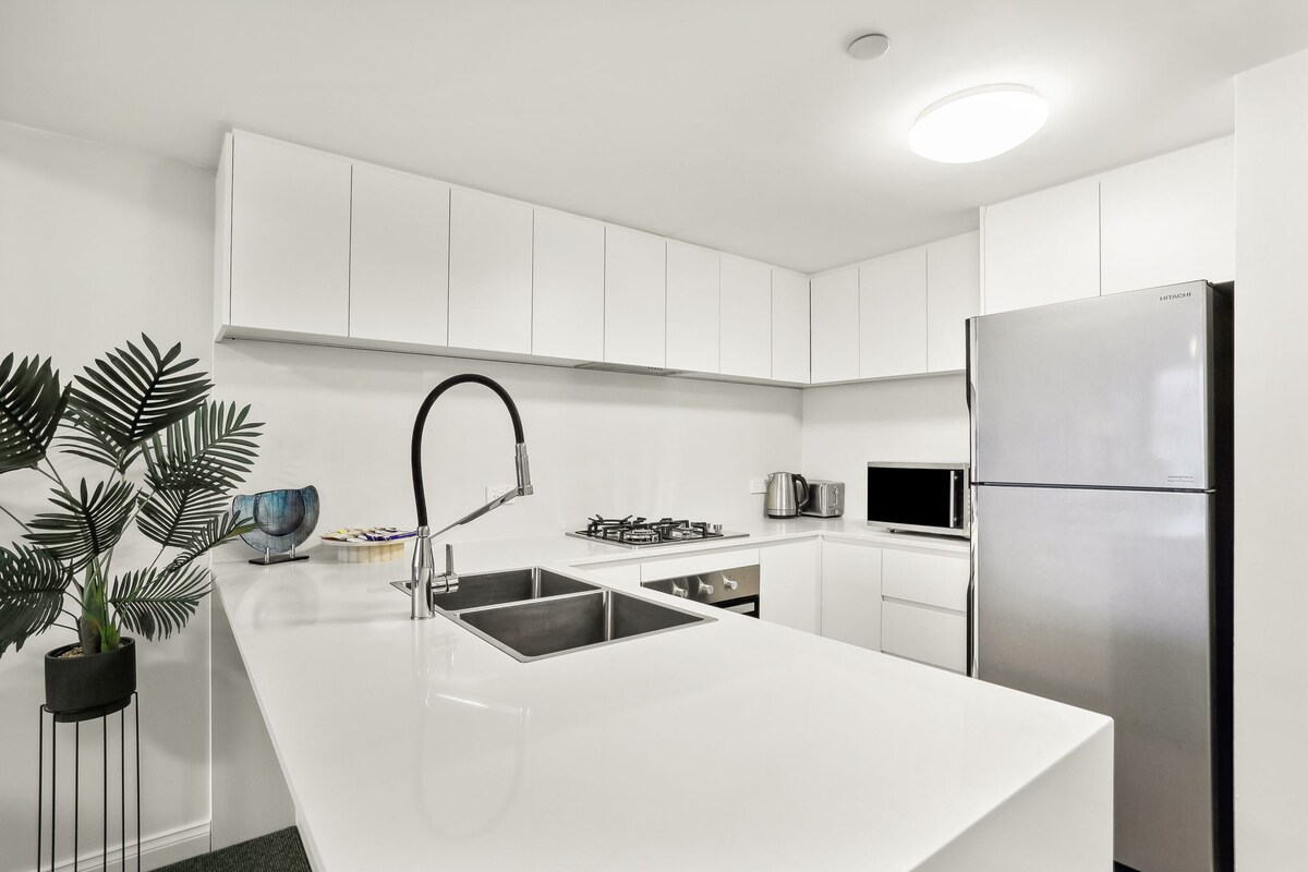 A modern kitchen features sleek white cabinetry complemented by stainless steel appliances, including a refrigerator and microwave. A gas cooktop is positioned on the countertop, which is adorned with a polished stone surface. A single basin sink and a decorative plant add aesthetic appeal.