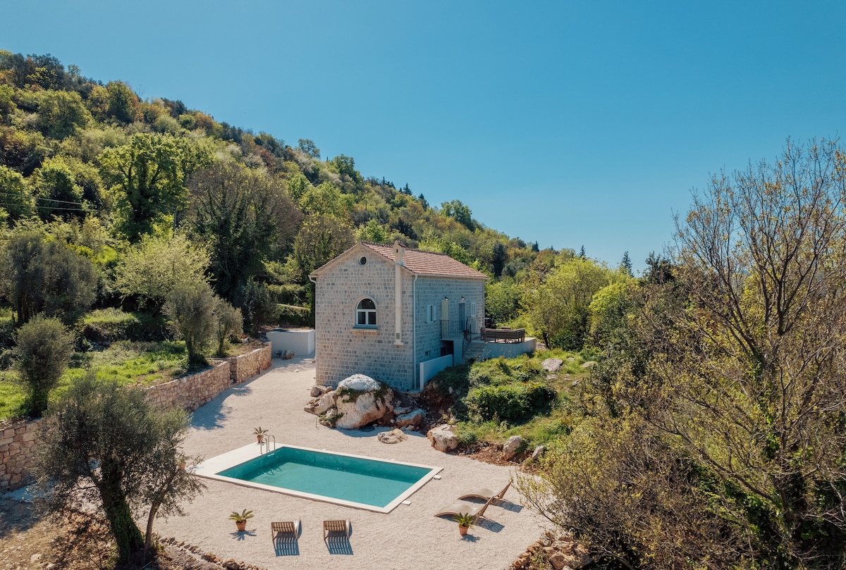 The house is set amidst lush greenery, with a stone exterior and a terracotta roof. A pool is situated in front, accompanied by sun loungers. The surrounding landscape features a variety of trees and natural rock formations, providing a serene atmosphere.