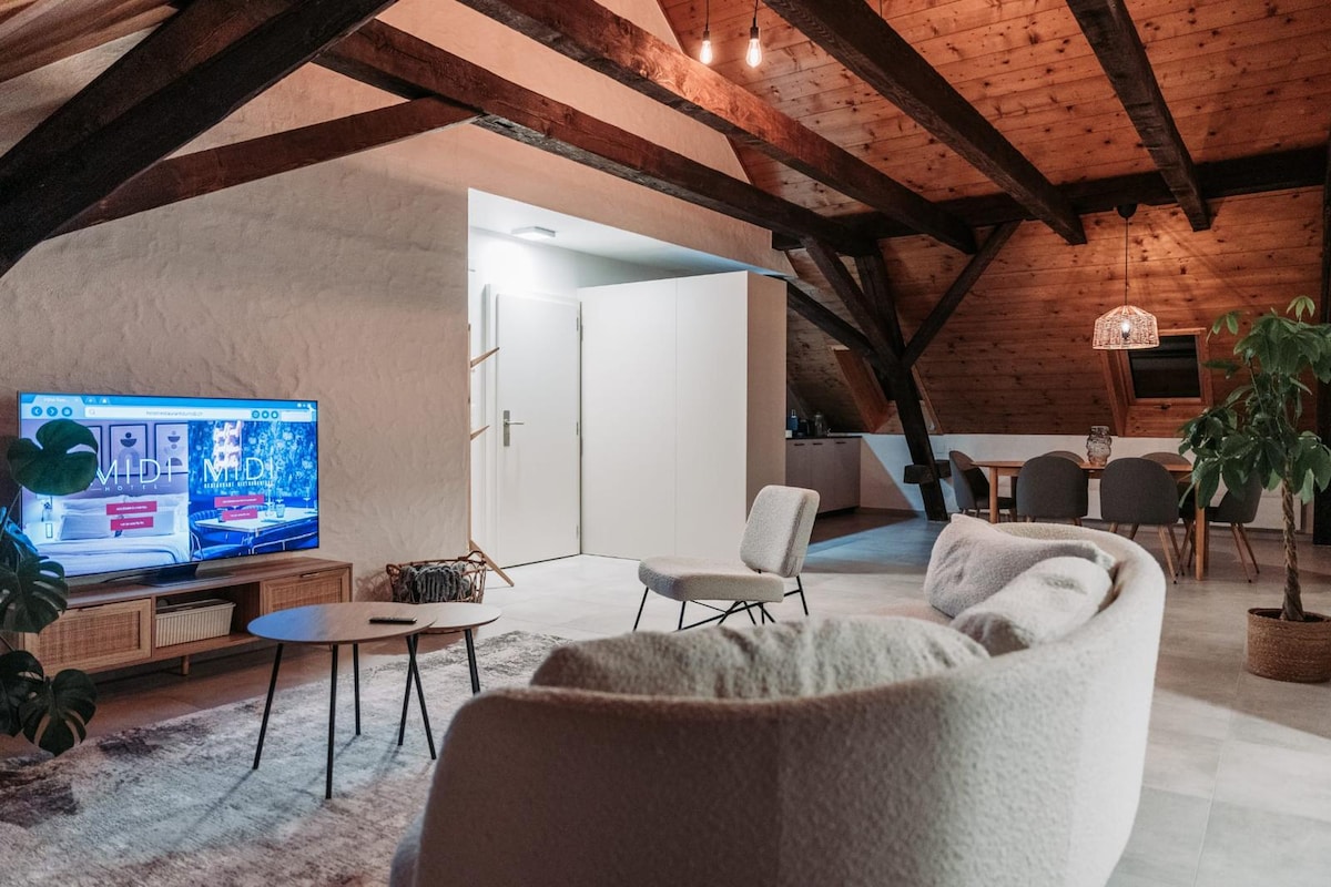 An open living area features a comfortable white sofa and a stylish wooden coffee table. A large television is positioned against the wall, with a cozy armchair near it. Natural wood beams highlight the ceiling, and a dining area is visible in the background.