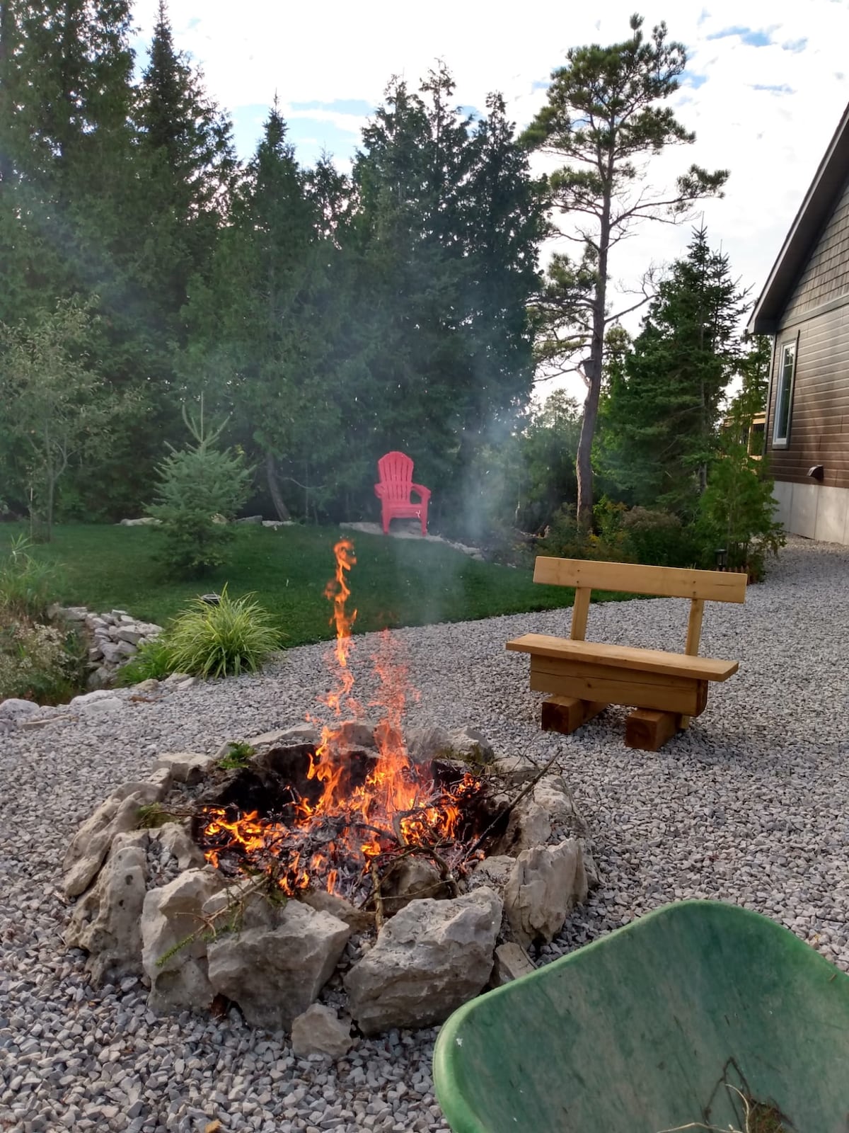 A fire pit constructed with stones is depicted, with vibrant flames rising amid smoky air. A wooden bench sits nearby, providing a resting place. In the background, a bright red chair stands among greenery, illustrating a calm natural setting.
