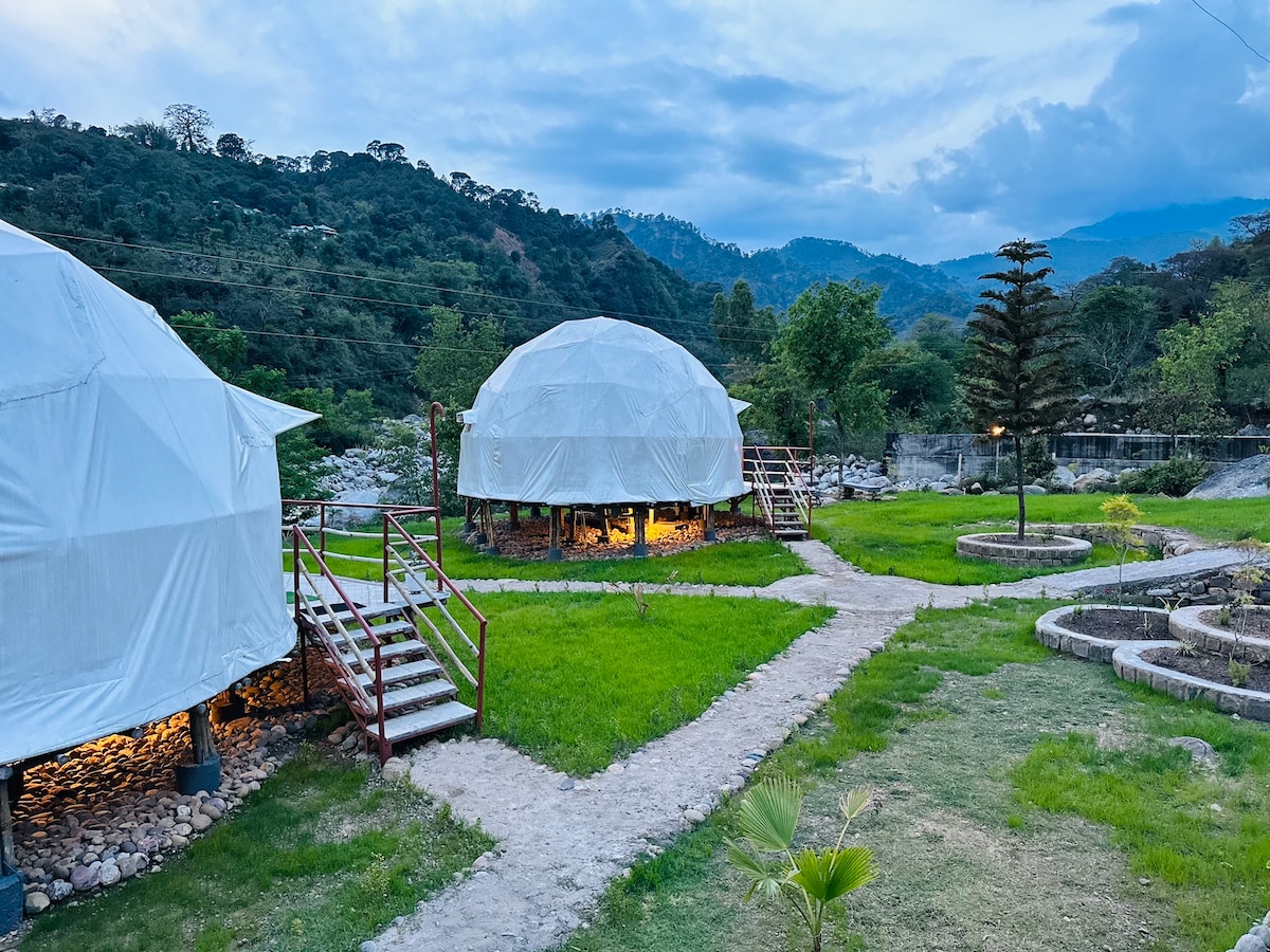 Two geodesic domes are nestled in a grassy area, surrounded by trees and mountains in the background. Pathways made of cobblestones and grass lead to each dome, while soft lighting from beneath the domes adds a gentle glow to the serene setting.