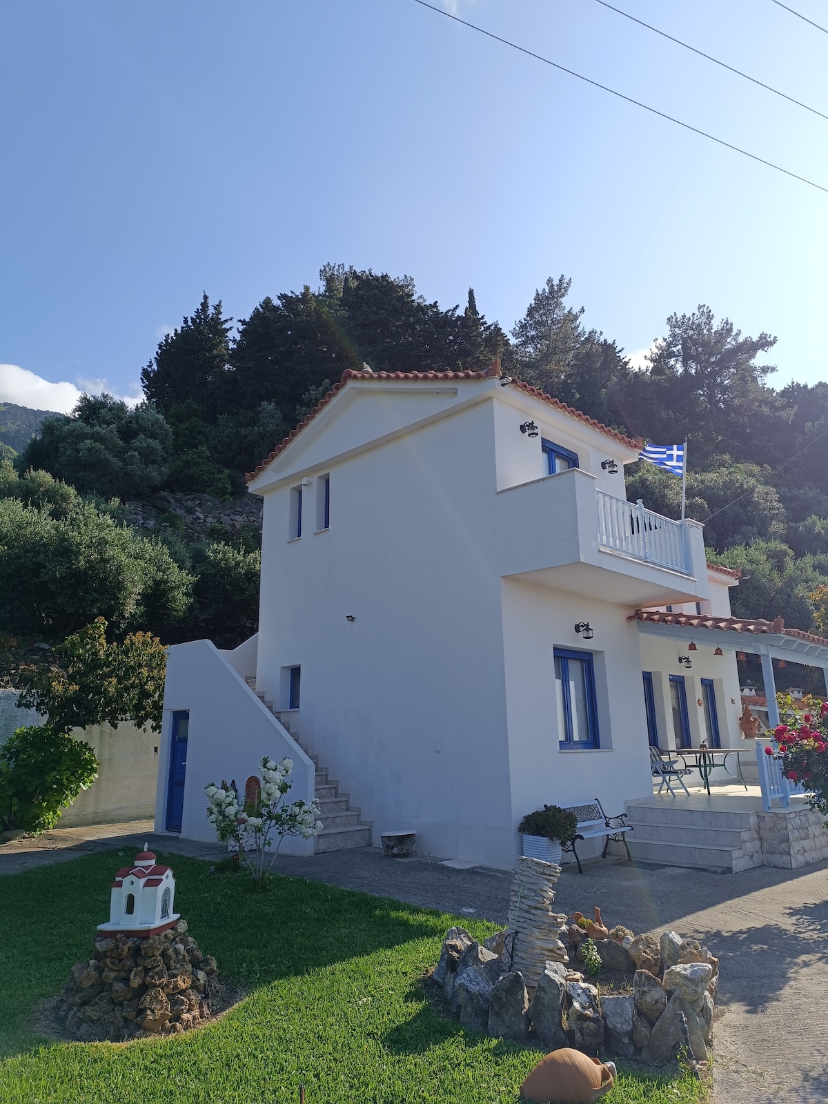 A two-story house with a white exterior and blue accents is shown, surrounded by greenery. A staircase leads to the entrance, while a small decorative structure is visible in the foreground. A flag is displayed on the upper balcony against a clear sky.