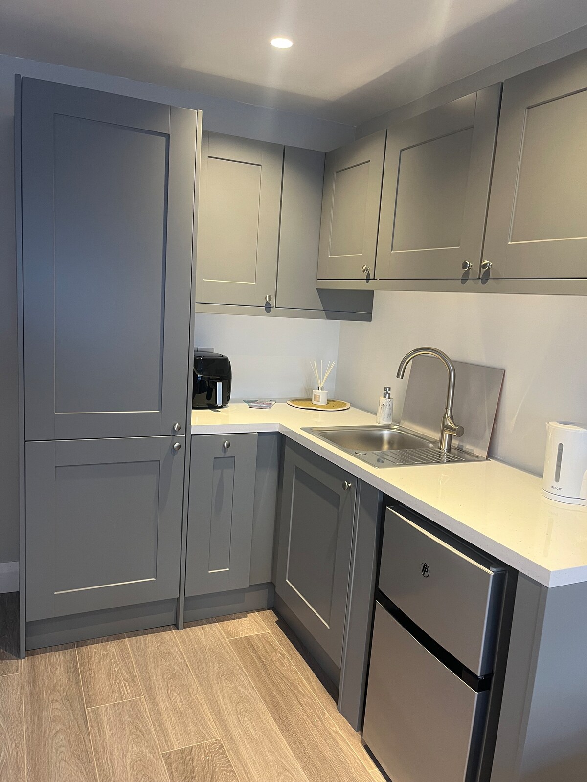 A modern kitchenette features grey cabinetry and a sleek countertop. Included are essential appliances such as a coffee maker, a sink, and a refrigerator. Simple decor and neutral tones create a functional space, ideal for preparing meals.