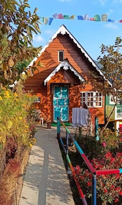 A charming wooden cottage is framed by vibrant flower beds and greenery. The exterior showcases a sloped roof adorned with white accents and colorful prayer flags fluttering above. A pathway leads to a bright blue door, inviting guests into a warm and welcoming space.