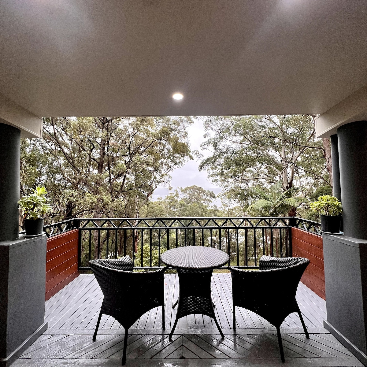 A private balcony is presented with a round table and two wicker chairs, surrounded by lush greenery. Potted plants add a touch of nature, while the quiet atmosphere suggests a peaceful outdoor space for relaxation. The wooden decking complements the natural surroundings.