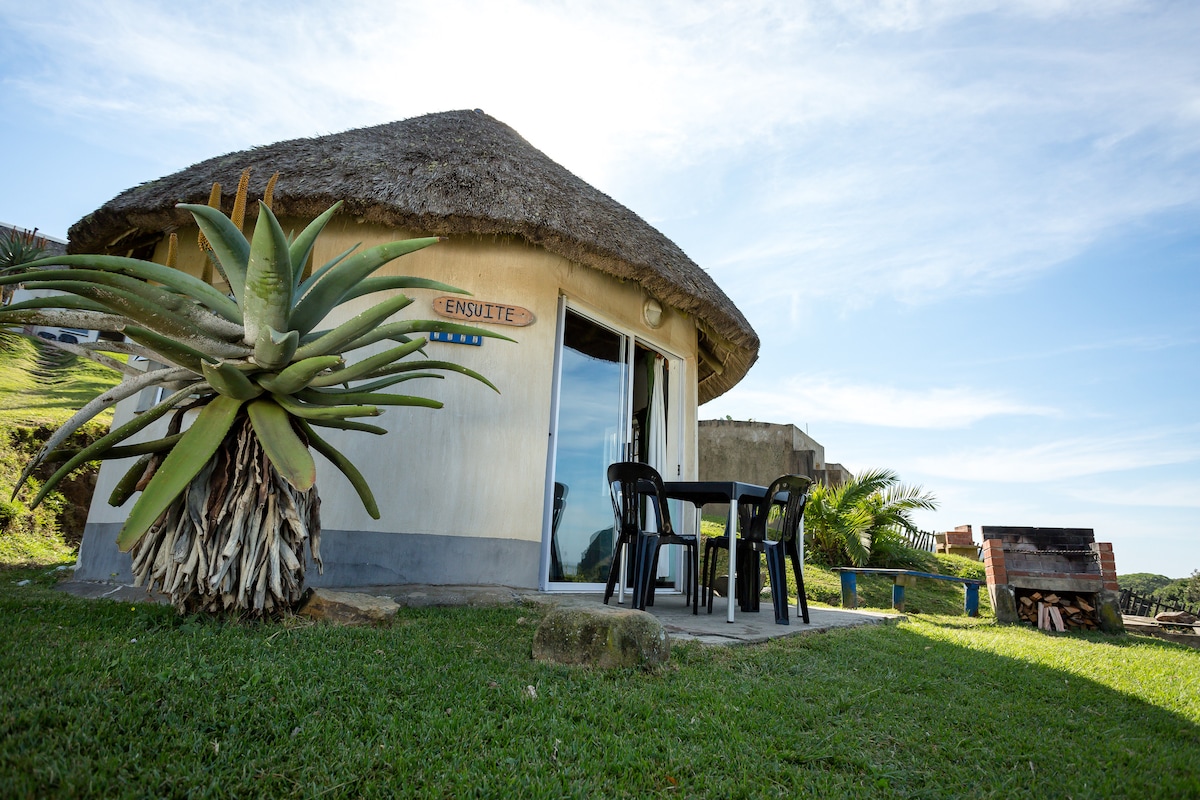 A round, thatched-roof structure is shown, with large glass doors reflecting natural light. Two outdoor chairs and a small table are positioned on the grassy area in front, offering a serene view of the surroundings.