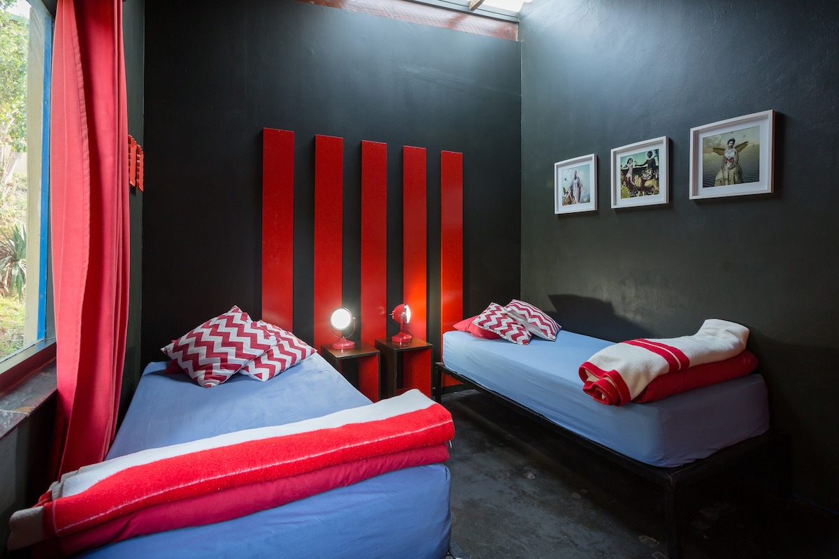 The room features two single beds dressed in blue linen, flanked by bedside tables with reading lamps. A large window allows natural light to enter, while red curtains frame the glass. The dark walls are complemented by bright red accent panels and framed photographs.