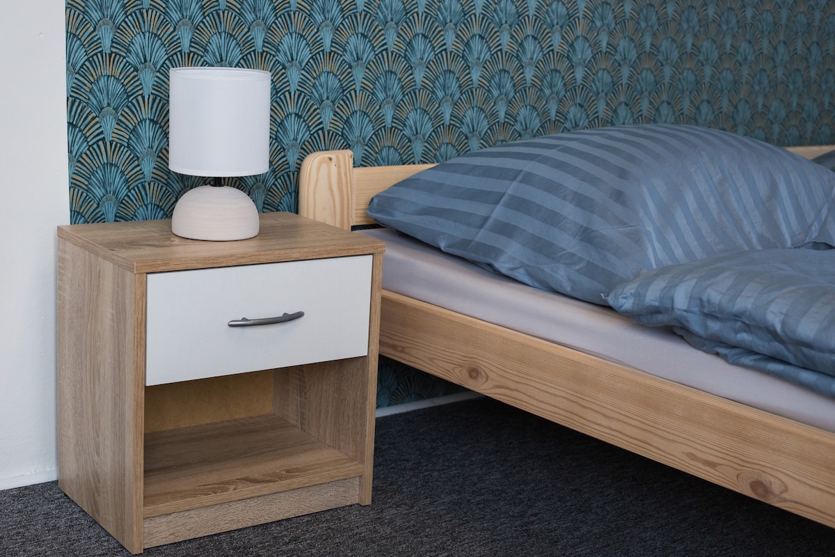 A bedroom corner features a light wooden bed with striped bedding. A bedside table with a single drawer stands alongside the bed, topped with a small lamp. The wall is adorned with a patterned blue wallpaper, adding a subtle design element.