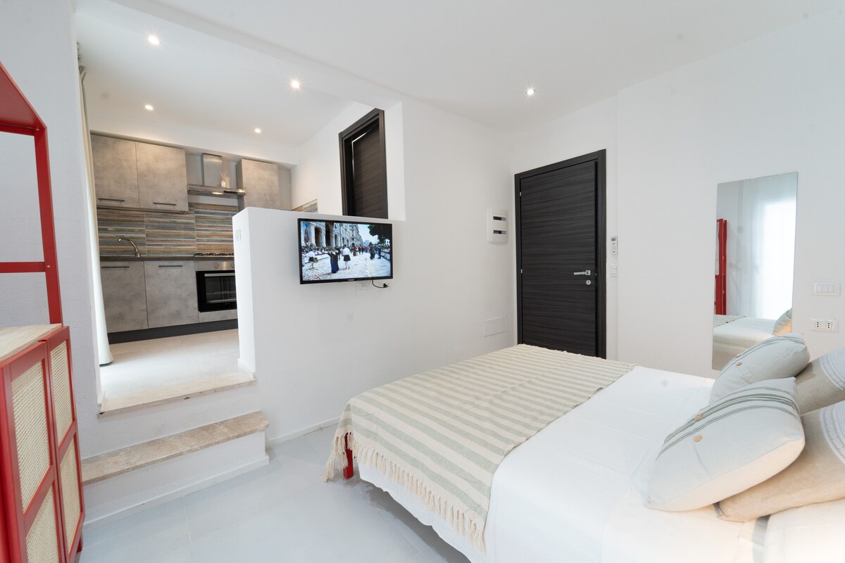 A well-lit bedroom features a spacious bed with a striped blanket and decorative cushions. A mounted smart TV displays a scenic image. A mirror reflects the room's modern design, while a kitchenette area is visible in the background.