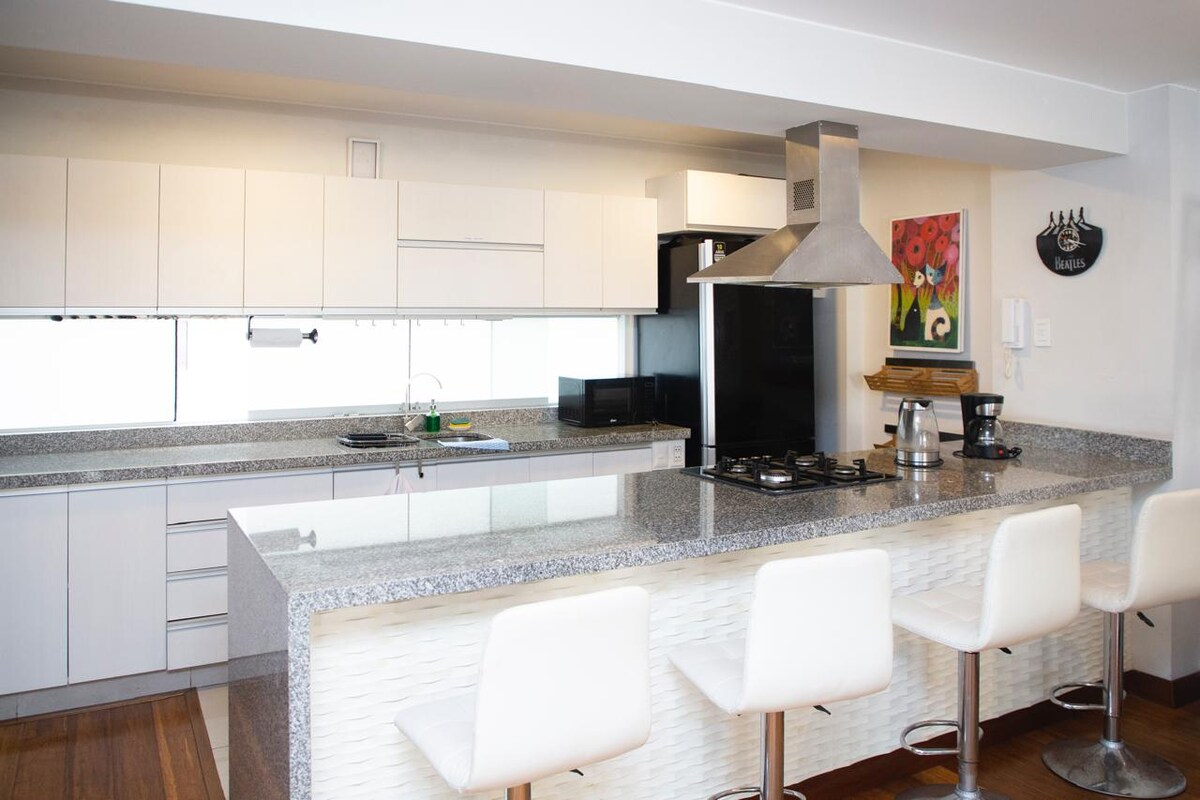 The kitchen features a spacious countertop with a modern design, complemented by sleek white cabinets. A gas stove and coffee maker are visible, alongside barstool seating. Light streams in through the large window, offering a bright and airy cooking environment.