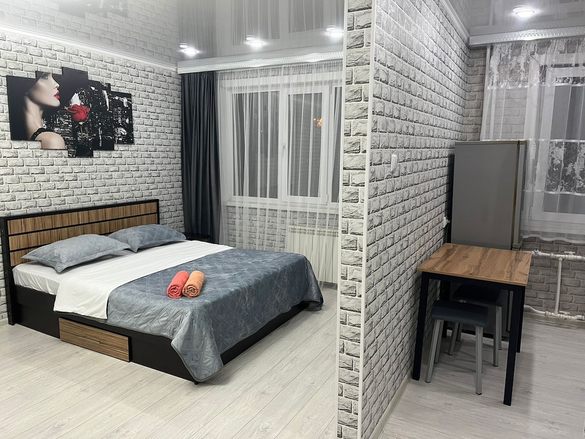 A bedroom is presented with a spacious bed dressed in grey linens and accented by colorful towels. A small, simple dining table with chairs is positioned nearby. Natural light enters through a window adorned with sheer curtains, and textured white brick wallpaper enhances the space.