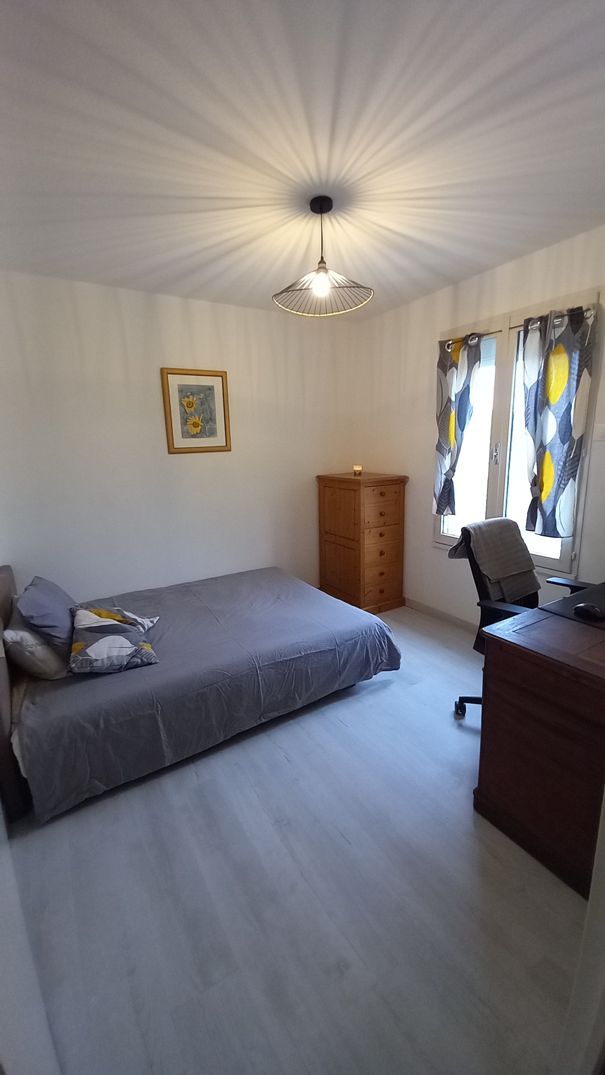 A well-lit bedroom features a spacious bed layered with a light grey cover. A wooden dresser stands against one wall, while a desk with a chair is positioned nearby. Curtains with a modern pattern frame the window, and a decorative wall piece adds a touch of color.