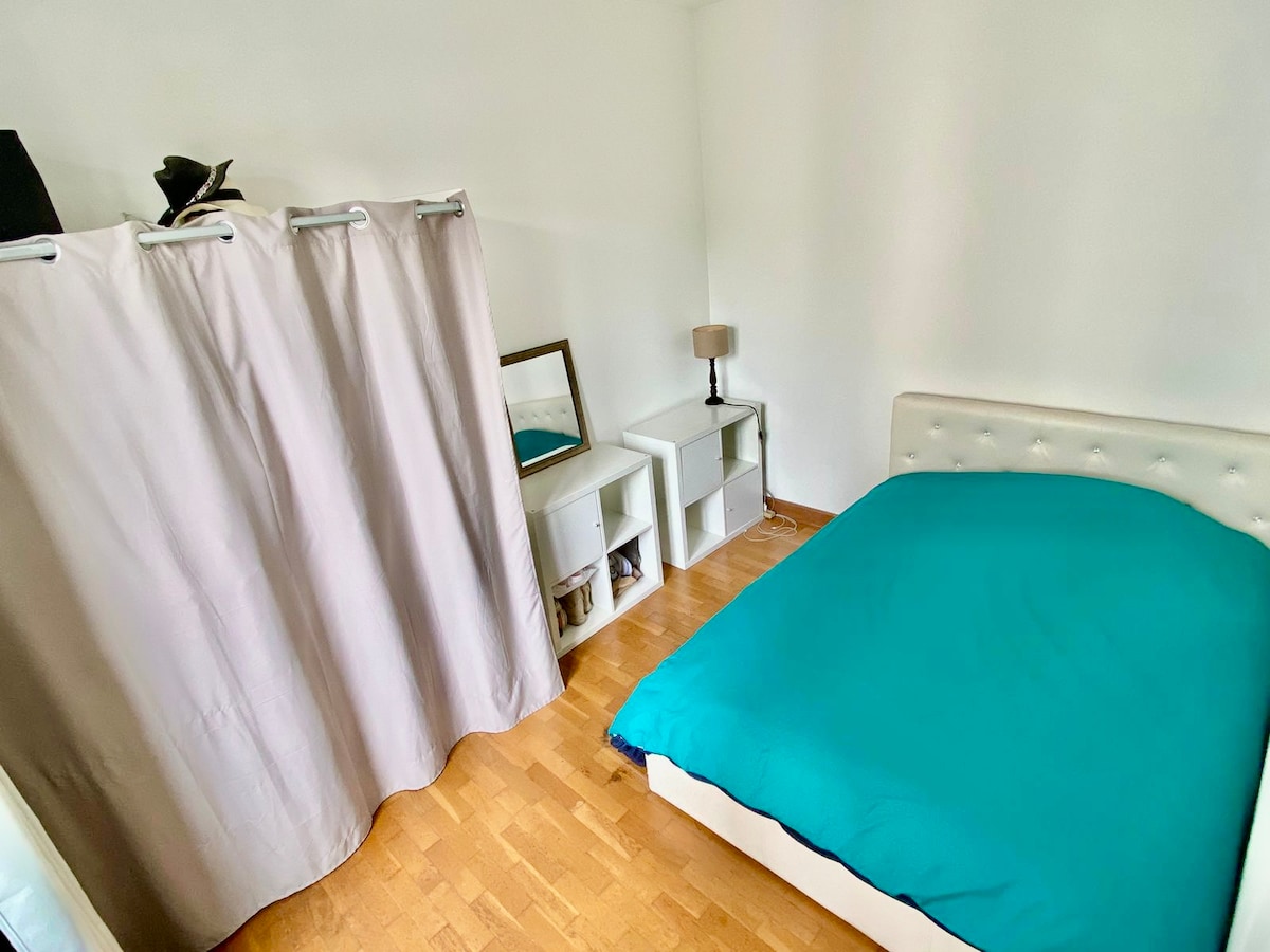 A private sleeping area features a spacious bed with a bright turquoise cover. A light-colored curtain partially divides the room, adding privacy. A small bedside lamp sits beside the bed, and minimalist shelving is visible against the wall.