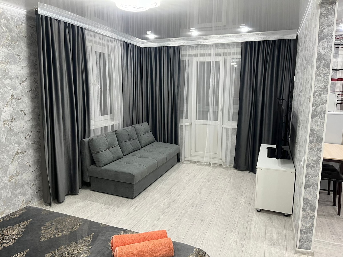 A comfortably furnished living area features a gray sofa against a light-colored wall. Large windows dressed with sheer curtains allow natural light to fill the space. A neutral-toned floor complements the overall décor, while a television is positioned on a sleek white stand.