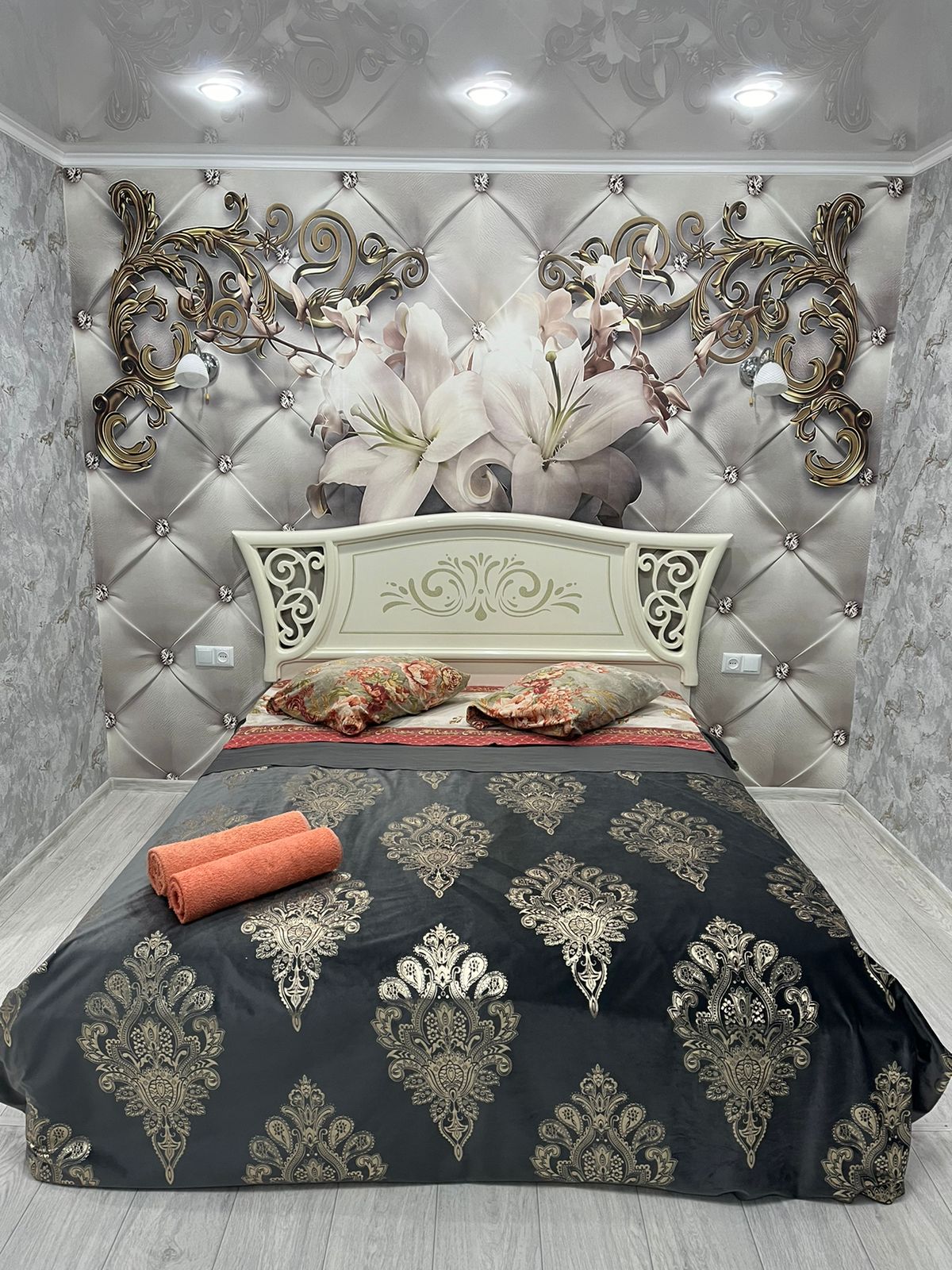A well-appointed bedroom features an ornate headboard against a floral wall mural. The spacious bed is dressed with decorative pillows and a patterned coverlet. Soft lighting from the ceiling highlights the elegant design, while rolled towels are neatly placed at the foot of the bed.