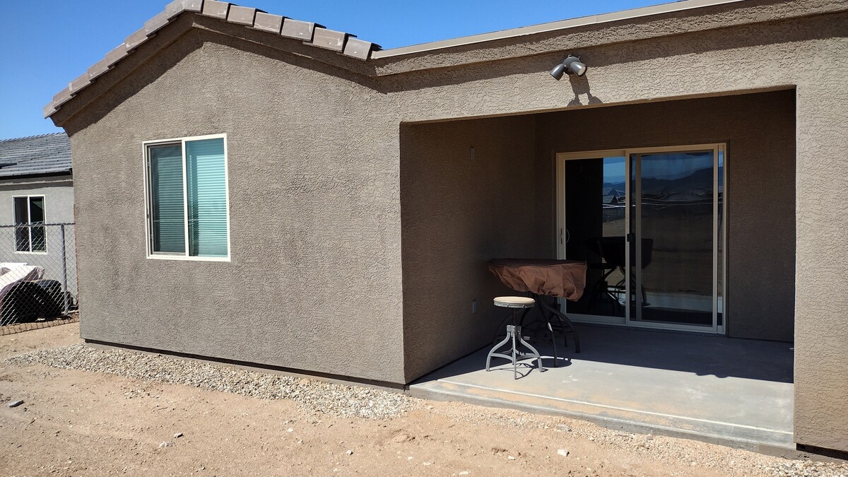 The exterior of a single-story home is shown, featuring a textured beige stucco facade. A spacious patio area is visible, with a round table and chairs set up beside sliding glass doors that lead into the home. A nearby window allows light into the interior.
