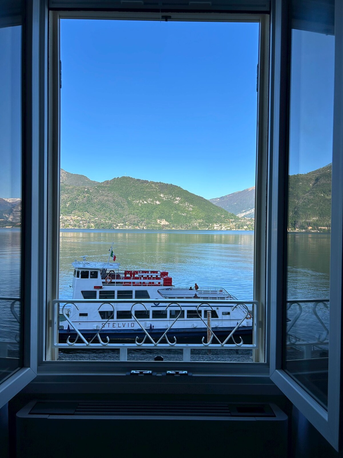 A serene view from the window reveals Lake Como, framed by opened glass panes. A ferry boat waits at the dock, with mountains rising majestically in the background against a clear blue sky.