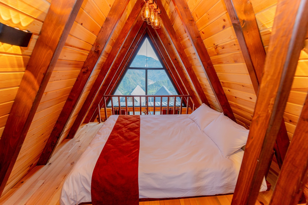 An inviting sleeping area features a spacious bed positioned beneath an a-frame ceiling. Natural wood beams and paneling create a warm atmosphere. A large window offers a view of the surrounding landscape, allowing natural light to illuminate the room.
