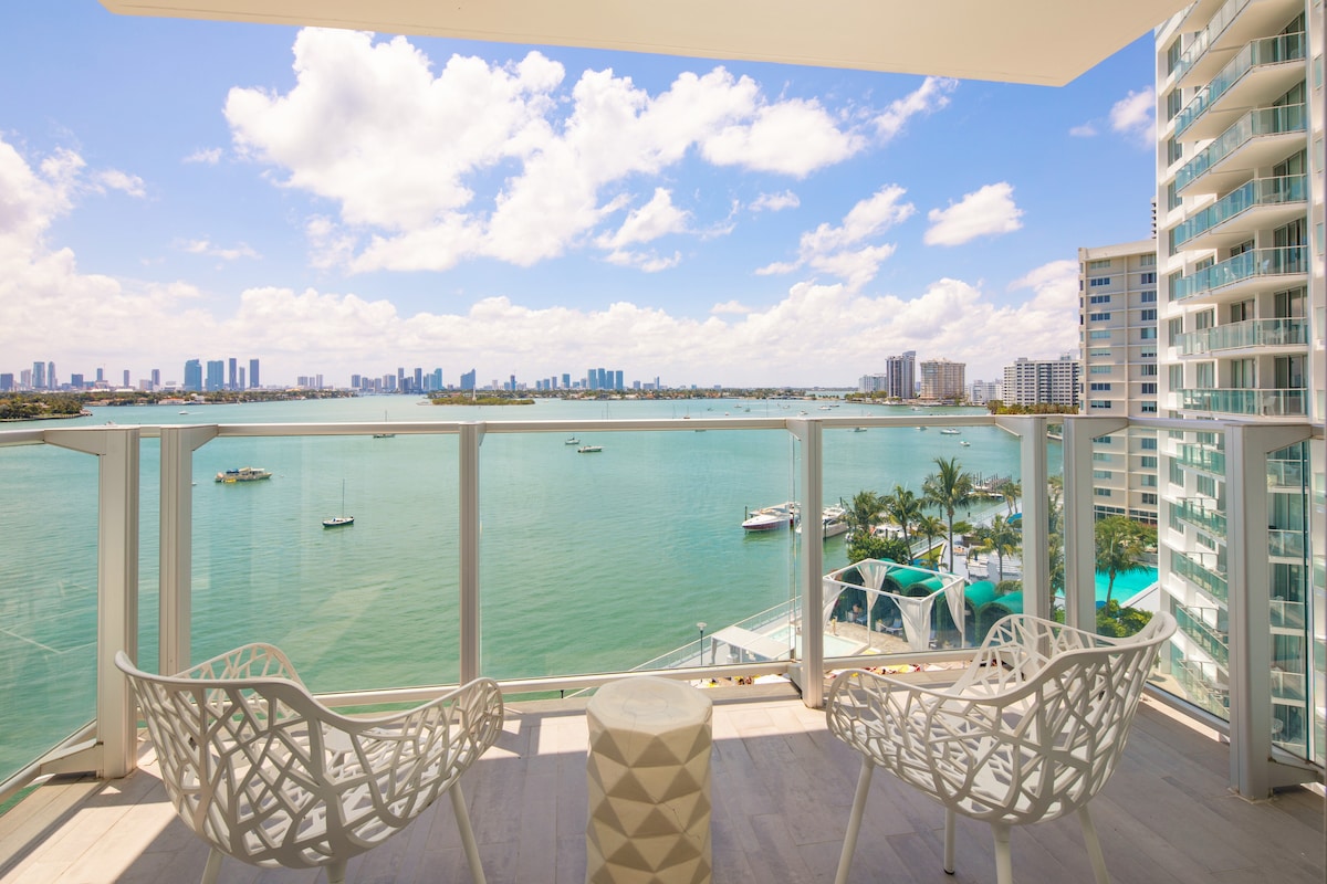 A spacious balcony features two intricately designed chairs facing the serene waters of Biscayne Bay. The expansive view includes distant city skyscrapers against a backdrop of fluffy clouds and a bright blue sky, creating an inviting outdoor space for relaxation.