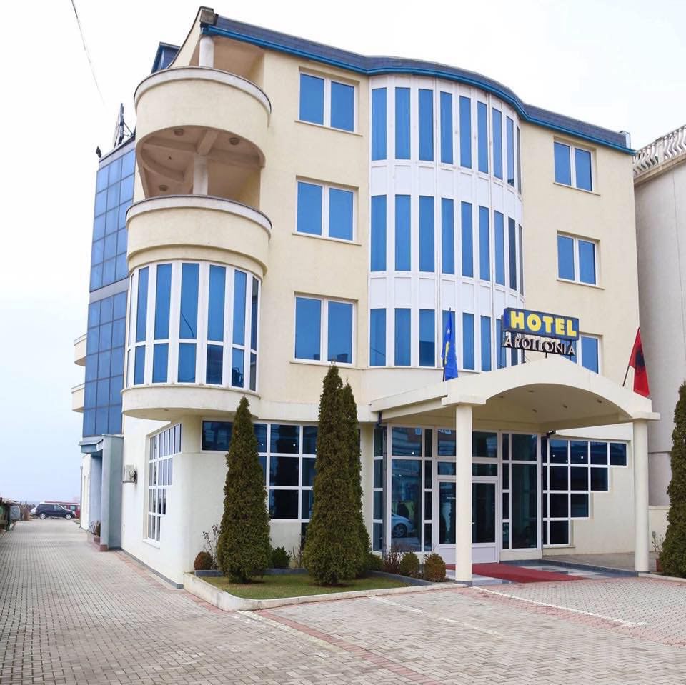 The hotel exterior showcases a modern three-story building with large glass windows and a pale yellow facade. Two well-trimmed trees flank the entrance, leading to a spacious lobby area. The surrounding paved area offers parking and clear visibility of the hotel signage.