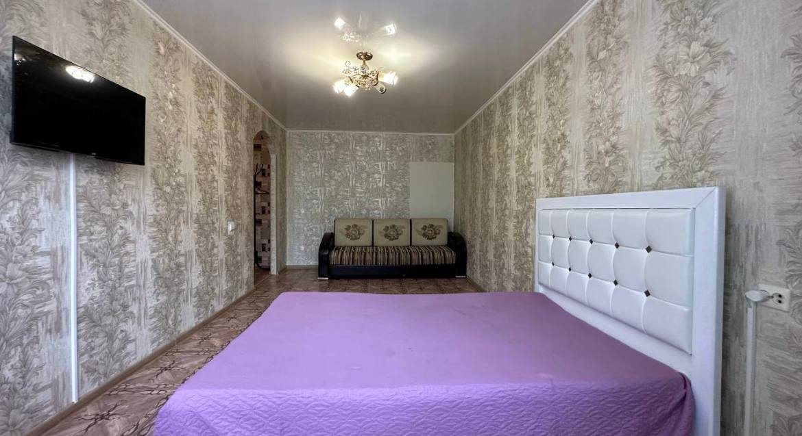 The spacious room features a light purple bedspread over a large bed, complemented by a dark sofa against the wall. A floral wallpaper creates a soft backdrop, while a flat-screen TV is mounted nearby, contributing to the room's functional design.