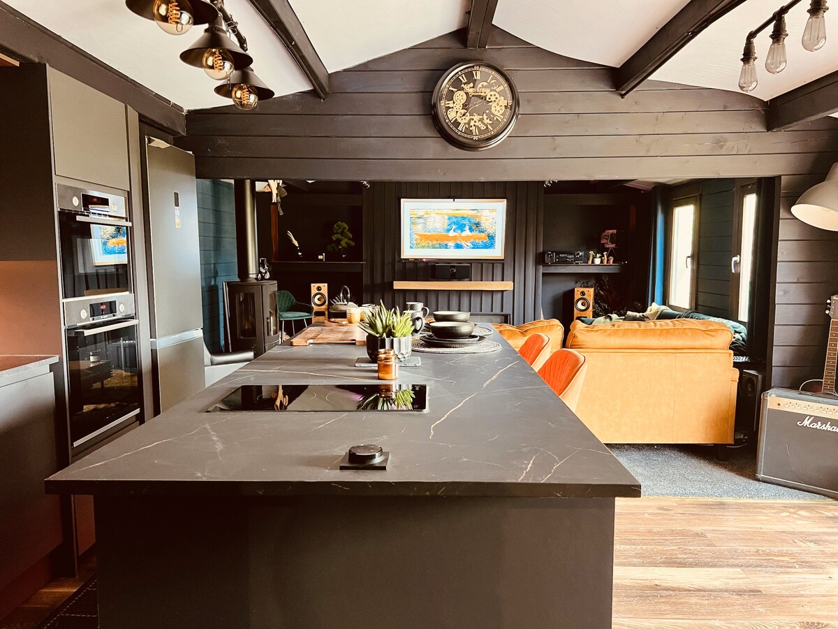A modern kitchen and living area is showcased, featuring an island with a dark surface and induction hob. The seating area includes orange chairs and a comfortable sofa. Artistic wall decor and soft lighting contribute to the stylish ambience, with large windows allowing natural light.