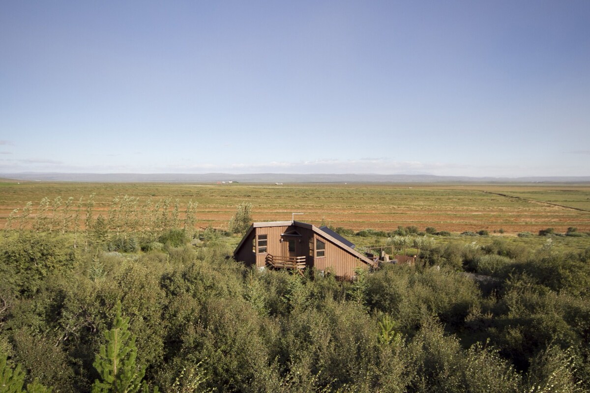 A wooden lodge is nestled among green foliage, surrounded by an expansive landscape. Rolling hills extend into the distance under a clear blue sky, providing a serene backdrop. The building features a sloping roof and a deck that invites outdoor relaxation.