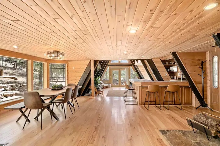 A spacious dining area features a wooden table surrounded by six chairs, set against a backdrop of natural wood walls and a light wood ceiling. Bar stools are positioned at the kitchen island, creating an open flow to the cozy living space filled with sunlight from expansive windows.