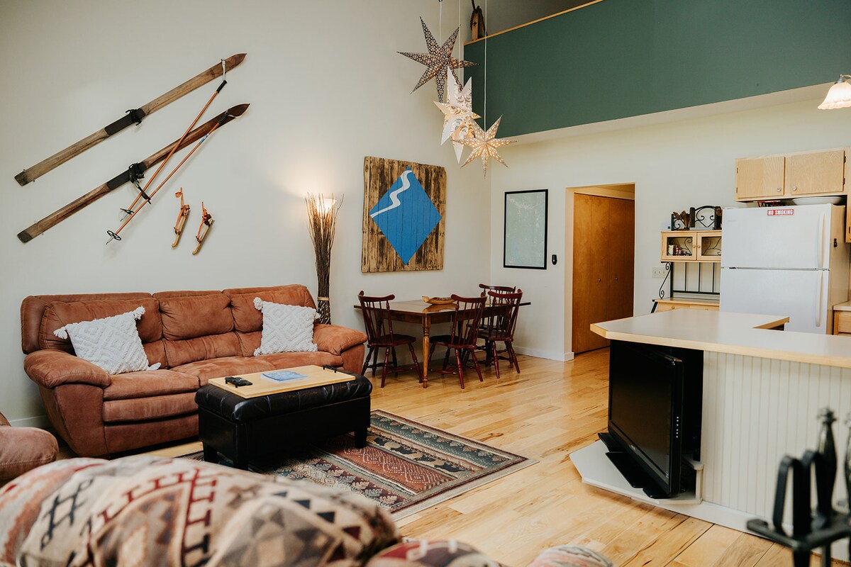 A spacious living area features a combination of rustic and modern decor. The room is equipped with a comfortable brown sectional sofa, a dark coffee table, and a patterned rug. A dining area with wooden chairs is visible, along with ski-themed decor and a well-equipped kitchen.