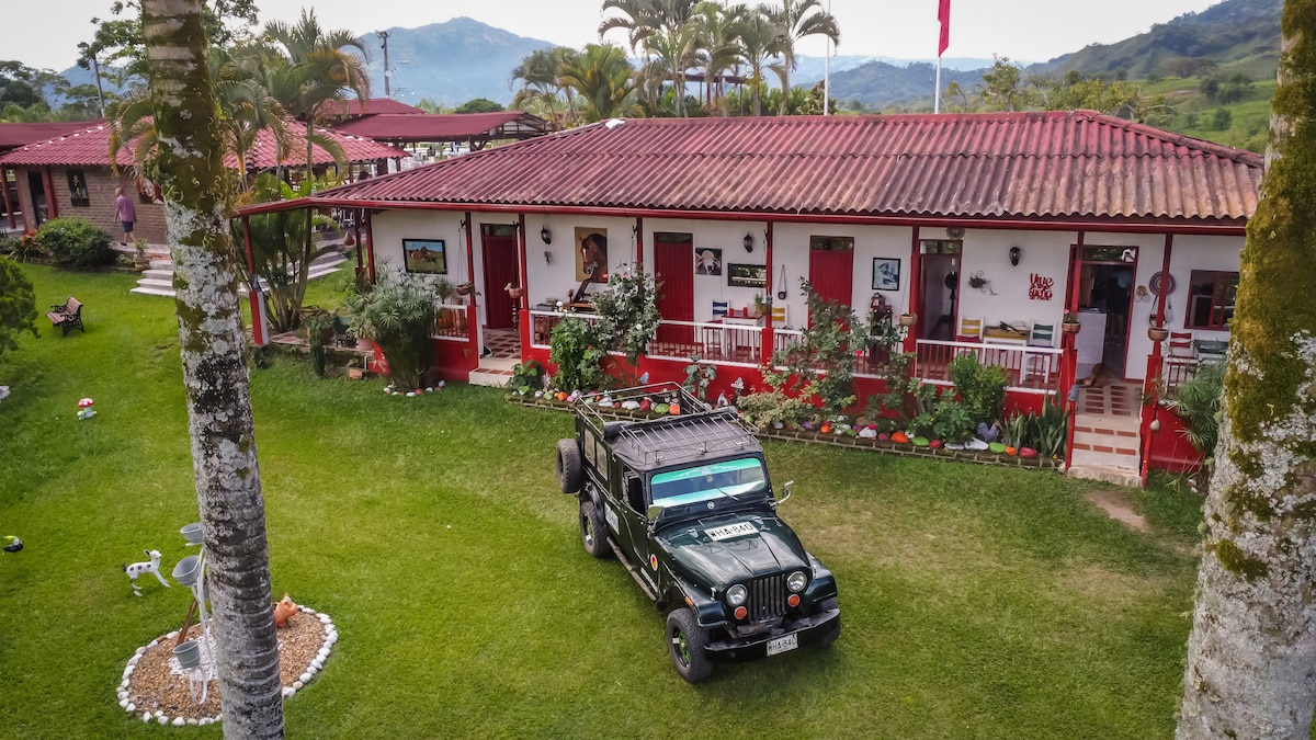 The exterior of a rustic building showcases a vibrant red and white color scheme, surrounded by lush greenery and tropical plants. A vintage jeep is parked on the grassy area in front, with open seating areas and decorative elements visible along the porch.