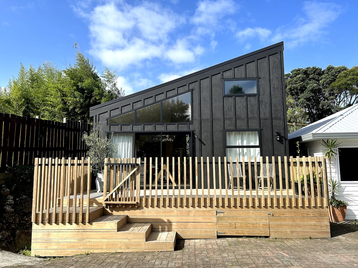 A modern black house features a sloped roof and large windows, bathed in natural light. A wooden deck with a railing surrounds the entrance, complemented by steps leading to the patio. Lush greenery is visible in the background, adding to the tranquil setting.