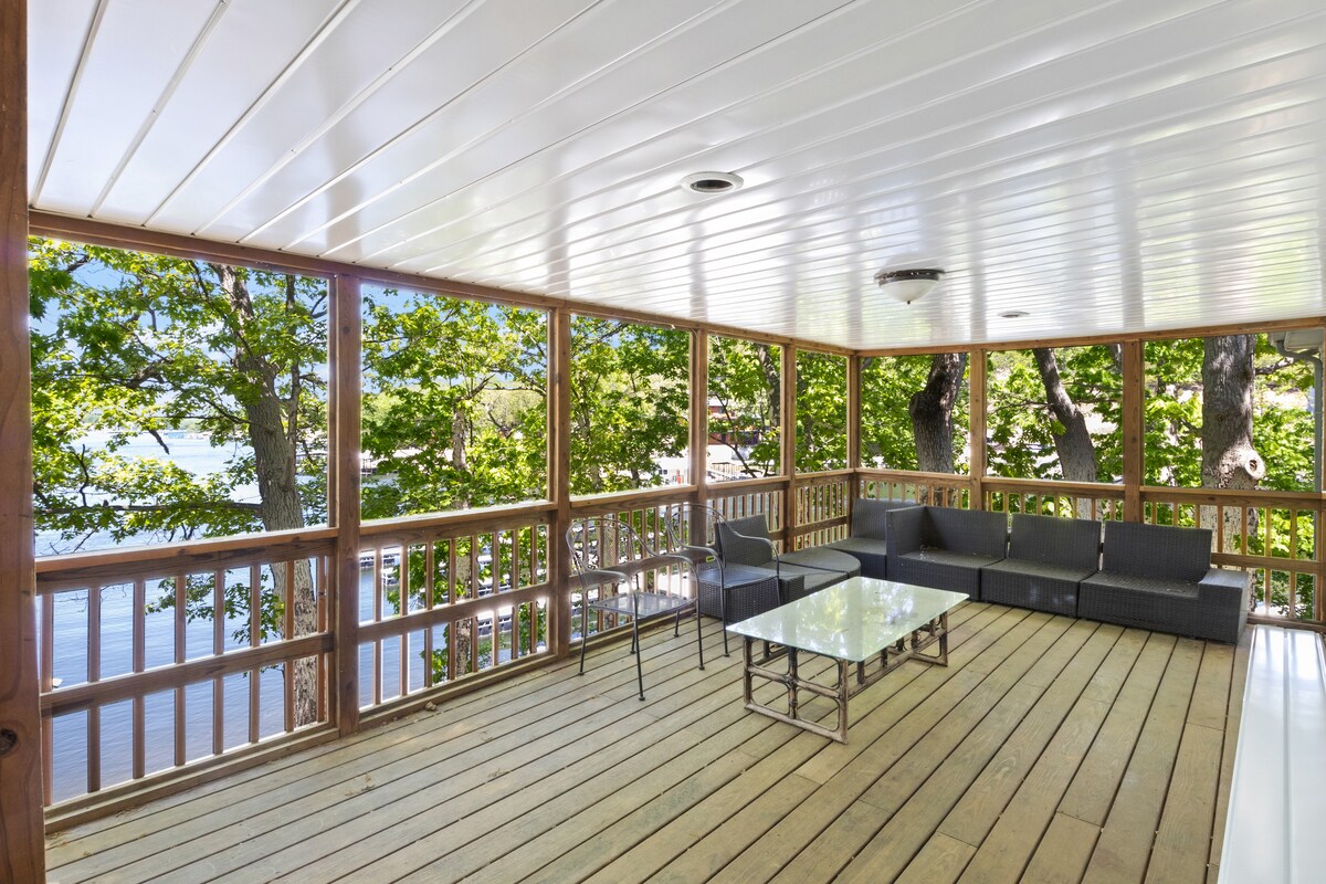 An expansive deck offers shaded seating with a modern sectional and a glass-top table. Views of the surrounding trees and water are visible through large open windows, enhancing the connection with nature. The wooden floor adds warmth to the inviting space.
