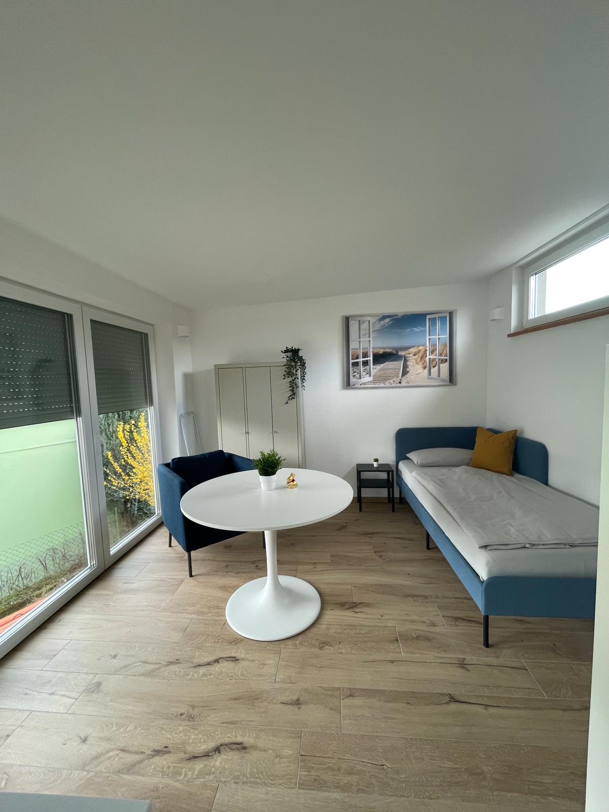 A bright living space features a light wood floor and large windows allowing natural light to fill the room. A small round dining table with a white base is surrounded by two chairs. A single bed is positioned against the wall, complemented by a yellow accent pillow.