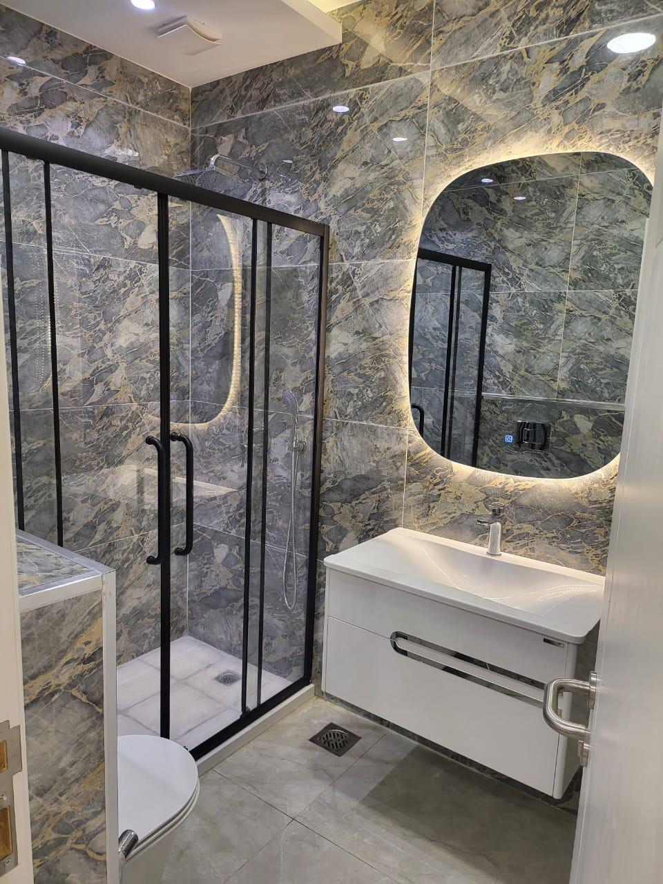 A modern bathroom features sleek stone walls and an elegant curved mirror. A spacious walk-in shower with glass panels is complemented by a minimalist sink area. Subtle lighting highlights the fixtures, creating a clean and inviting space.
