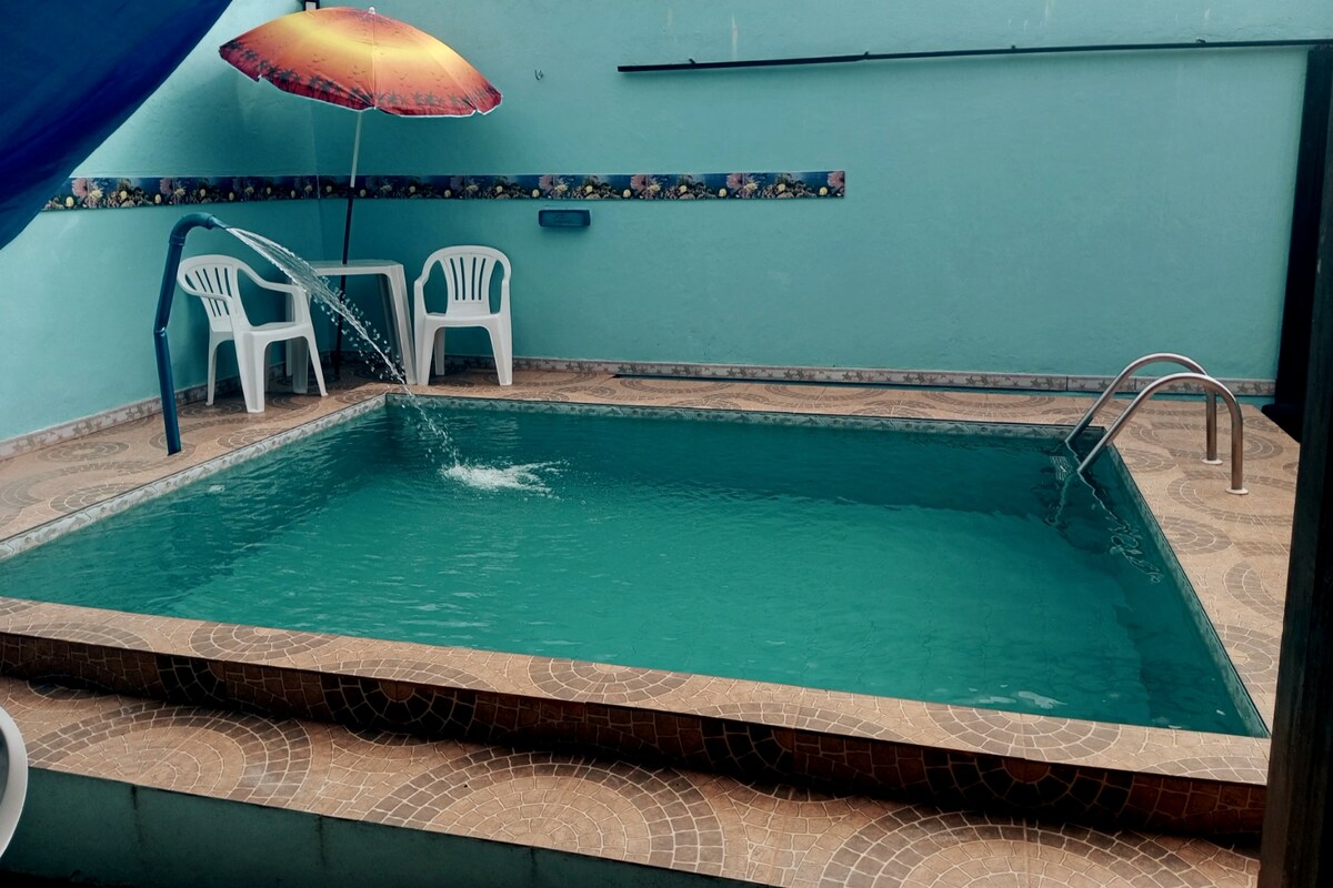 A turquoise pool is centered in a sunlit area, with water gently flowing from a spout. Two white chairs and an umbrella offer shaded seating. The pool area is bordered by textured tiles, and a decorative border enhances the surroundings.
