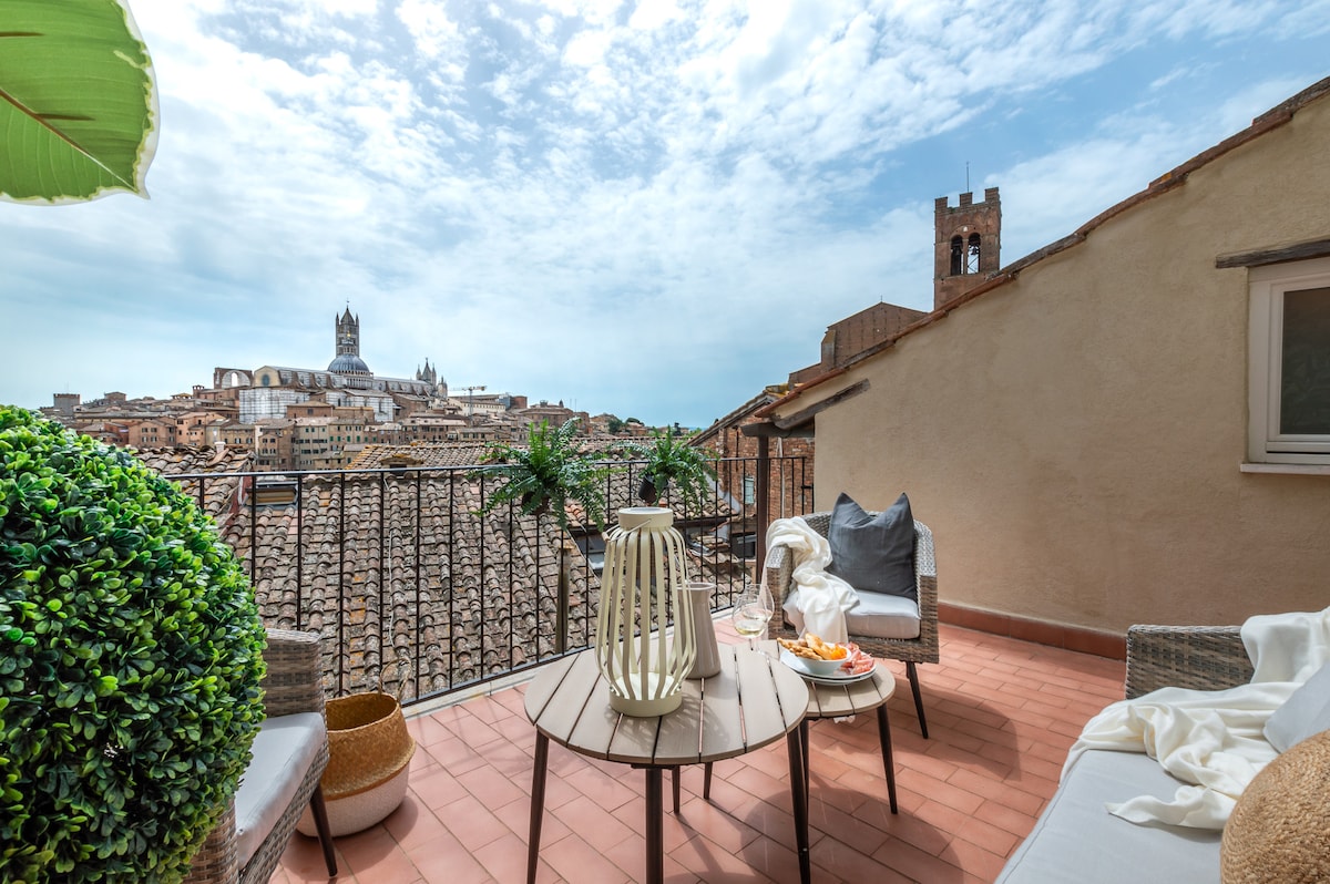 A terrace offers a spectacular view of Siena, showcasing the city rooftops and the Duomo in the distance. The outdoor seating area features a round table, two comfortable chairs, and decorative greenery. Natural light enhances the bright and airy ambiance, inviting relaxation.