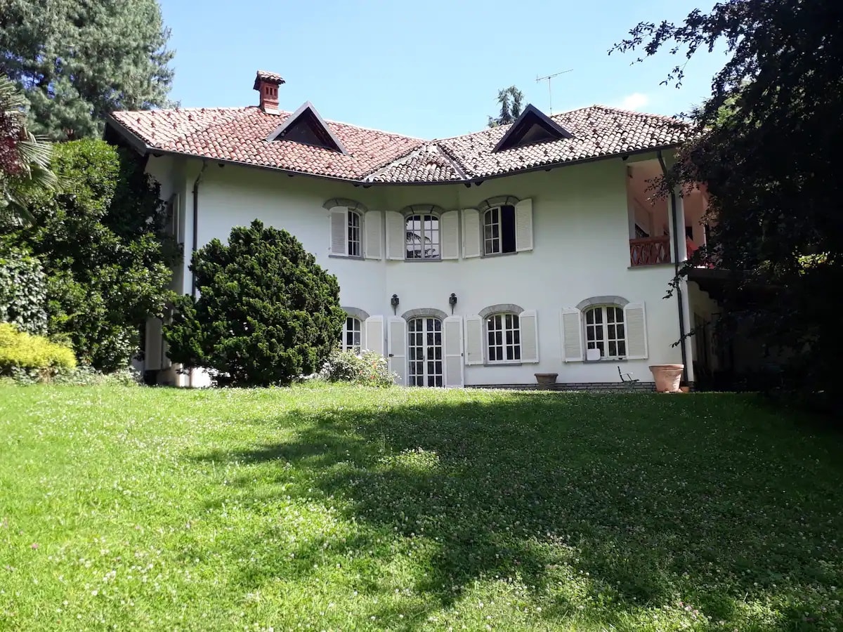 The villa's exterior is framed by lush greenery, featuring a white façade and traditional tile roof. Large windows with shutters provide a welcoming appearance. A well-kept lawn stretches in front, emphasizing the spaciousness of the property and the natural surroundings.