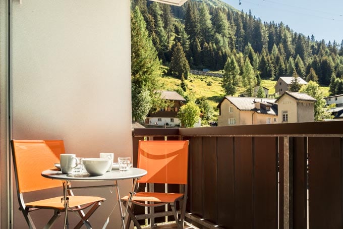 A balcony features two orange chairs surrounding a small round table set with dishes. The backdrop displays lush green mountains and charming village houses nestled among the trees, enhancing the outdoor space.