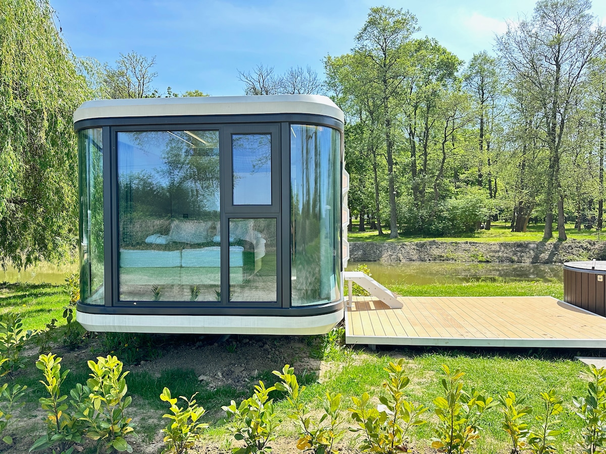 A modern glamping cabin features large glass windows, allowing ample natural light to brighten the inviting interior. A private deck extends towards a serene body of water, surrounded by greenery and trees, offering a peaceful retreat in nature.